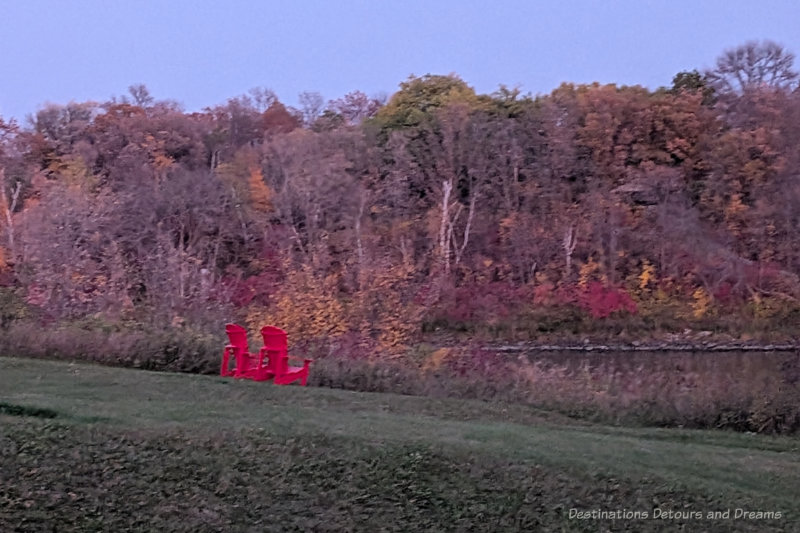 Red chairs along a river bank lines with trees with foliage of red and yellow fall colours