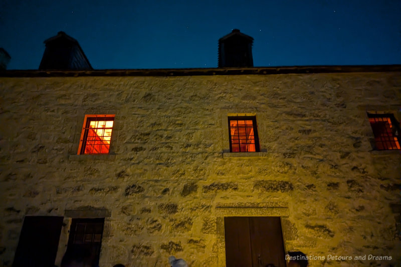 Upper outside wall of stone building at night with light shining through three windows