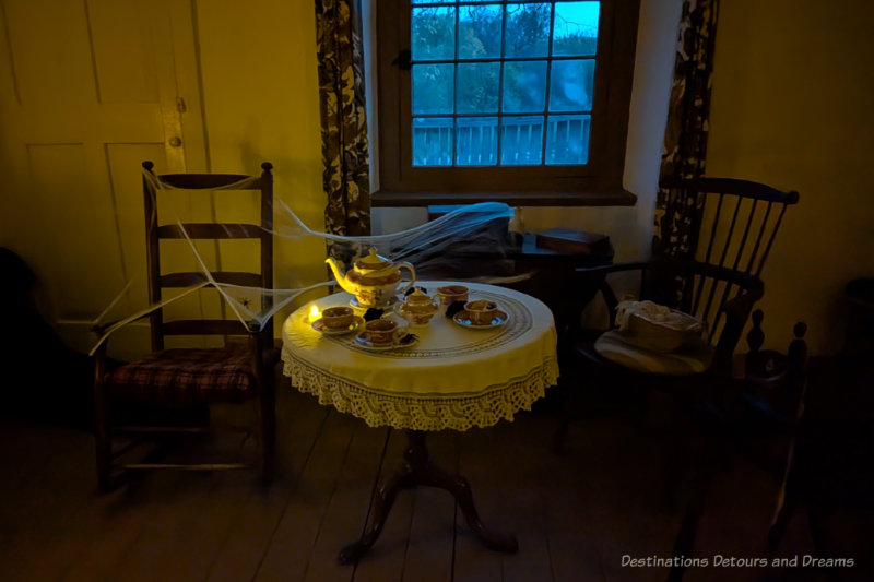 Circular table with white table cloth and teapot and tea cups as well as a couple of chairs covered in cobweb in dimly lit room